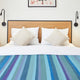Pululahua baby alpaca wool blanket – a bed featuring two white pillows and a vibrant, striped blanket in shades of blue. It is set in a simple room with a wooden headboard, wall sconces, and framed artwork
