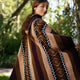 A person wears a colorful, patterned poncho featuring geometric designs and bold stripes, standing outdoors surrounded by lush green foliage and dappled sunlight.