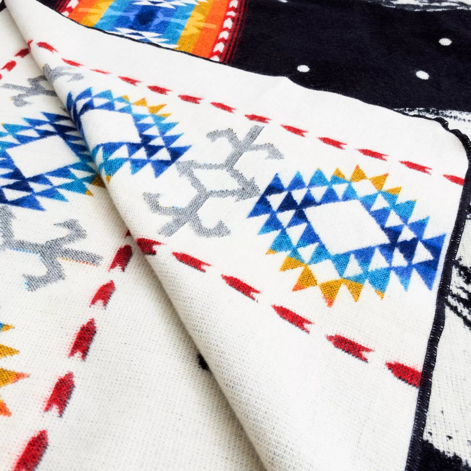 A colorful patterned textile lies folded, featuring geometric designs in blue, orange, and white, with red and gray accents. The background is a contrasting dark color.