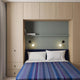 Morona baby alpaca wool blanket – bed with striped blue blanket, beneath a green wall shelf holding books, surrounded by light wood cabinets. Two wall lamps illuminate the bed, enhancing the modern bedroom setting