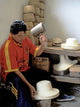 A person shapes a hat using a wooden mallet in a workshop. The room contains a workbench with stacked hats and boxes, indicating an artisan environment.