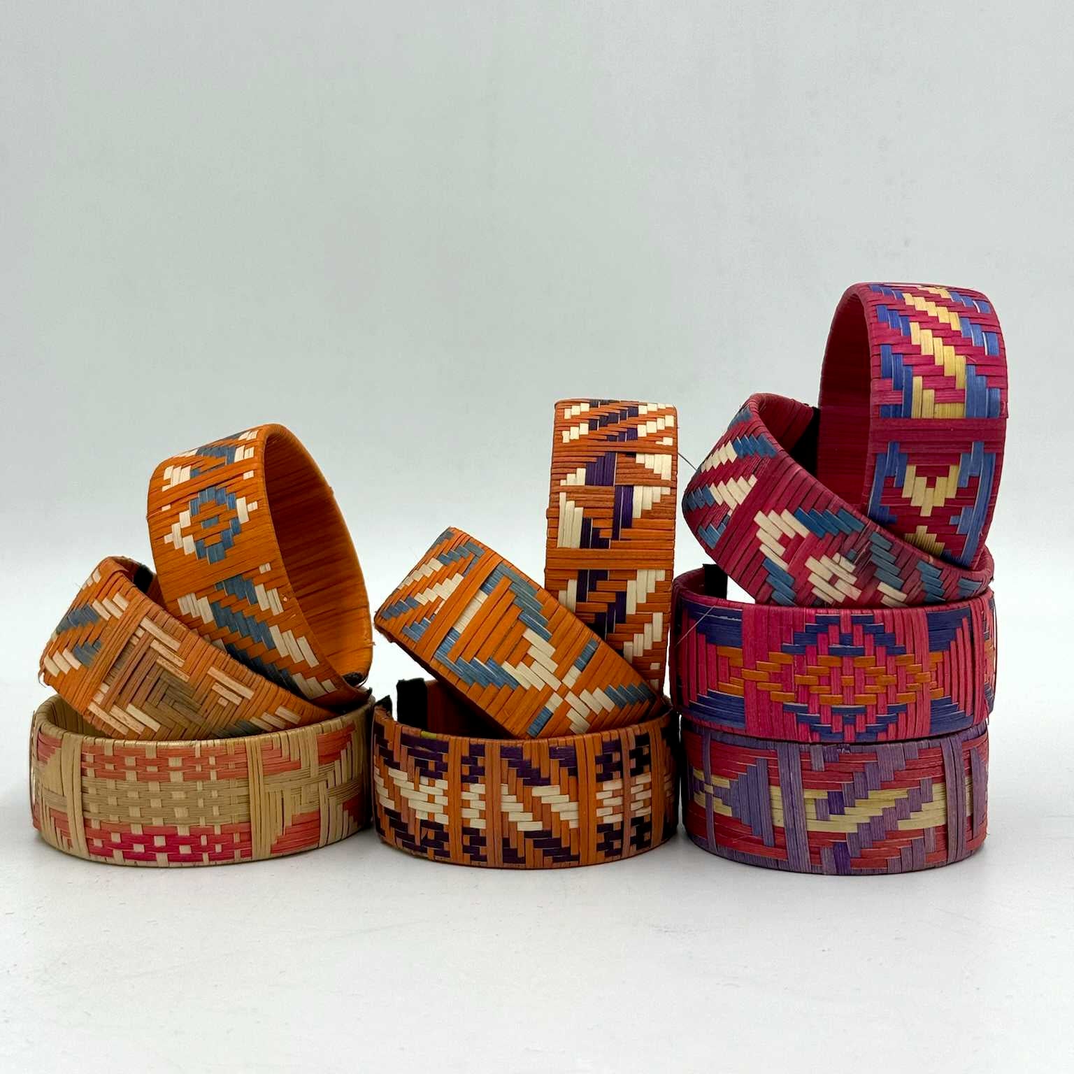 Colorful woven bracelets, featuring intricate geometric patterns, are stacked in two groups against a plain white background. The bracelets showcase vibrant orange, red, blue, and beige hues.