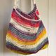 A crochet bag hangs, featuring colorful horizontal stripes of red, white, purple, green, yellow, and natural hues, set against a light-colored, vertical panel background.