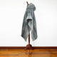 A gray poncho with intricate patterns is displayed on a wooden stand. The poncho drapes elegantly, showing its fringed edges, against a plain white wall and wooden floor.