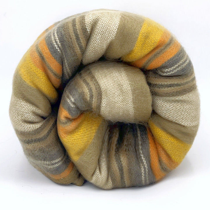 Rumi baby alpaca wool blanket – a rolled, knitted blanket with horizontal stripes in beige, yellow, and orange, tightly coiled. The background is plain white, emphasizing the blanket's colors and textures
