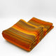 Cumanda baby alpaca wool blanket – a folded, striped blanket with warm colors like orange, yellow, and brown, lying on a plain white surface