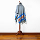 A gray poncho drapes gracefully over a wooden mannequin, featuring a colorful, patterned border with blue accents. It stands in a room with a wooden floor and plain white wall.