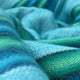 Machay baby alpaca wool blanket – a soft, woolen fabric with woven patterns undulates, featuring vibrant blue and green hues. The perspective is close-up, highlighting textures and gentle curves in a smooth, abstract environment