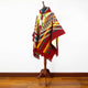 Guismi alpaca wool hooded poncho – a colorful poncho with geometric patterns displayed on a mannequin stand. It drapes elegantly, showcasing red, yellow, and green hues. The background features wooden flooring against a plain wall