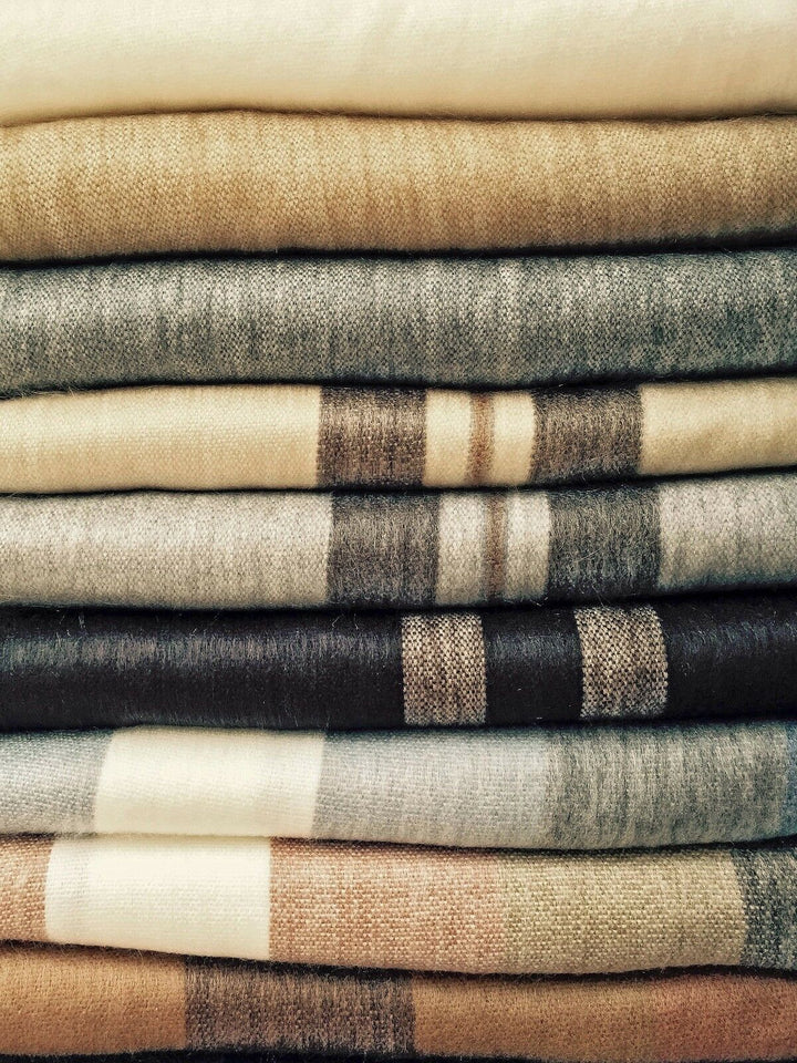 Wholesale lot of 30 queen alpaca wool blankets – stacked woolen blankets in shades of beige, gray, and brown, some with striped patterns, neatly arranged in horizontal layers, creating a textured and cozy appearance