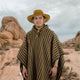 A person wearing a striped poncho and wide-brimmed hat stands in a rocky desert landscape with boulders and sparse vegetation under a cloudy sky.