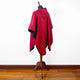 Red hooded cape displayed on a mannequin stand, with black trim, set against a plain white wall and wooden floor background.