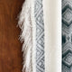 Cuyuja lightweight alpaca wool hooded poncho – a fringed, patterned gray and white poncho hangs vertically. The geometric design contrasts with a smooth wooden background, highlighting texture and color variations