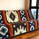 Qhusi extra large alpaca wool blanket – a colorful, patterned blanket drapes over a sofa in a modern room, featuring geometric designs in orange, blue, and black, near large windows overlooking a cityscape