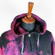 A wooden mannequin displays a black and pink tie-dye hooded sweater with a drawstring, against a plain white background.