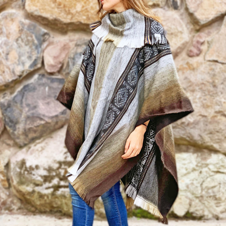 A person wears a patterned poncho with earthy tones, standing against a stone wall. The poncho features geometric designs and fringed edges. Jeans are visible underneath the garment.