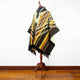 A patterned poncho featuring geometric designs in warm tones is displayed on a wooden mannequin. The setting is a room with wooden flooring and a plain white background.
