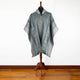Gray patterned poncho displayed on a wooden mannequin stand, set against a light-colored wall and wooden floor, highlighting its intricate design and fabric texture.
