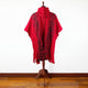 A red poncho, featuring black geometric patterns, drapes over a wooden mannequin stand. It is displayed in a room with a wooden floor and plain white wall background.
