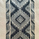 Thick, woolen fabric features a central diamond geometric pattern in dark gray, bordered by lighter bands, creating a textured, traditional design against a plain background.