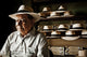 An elderly man wearing a white hat sits pensively. Behind him, shelves display similar hats in a dimly lit room. Boxes labeled Montecristi are visible on the shelves.