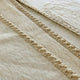 Beige wool blanket with textured, braided stitching runs lengthwise across the surface, highlighting a soft, consistent fabric pattern in close-up view.