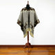 Poncho with geometric patterns drapes over a black mannequin on a wooden stand. The garment features gray fabric with a brown border, accented with tan fringe, against a white background.