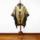A poncho with geometric patterns hangs on a wooden mannequin. It features earthy tones like brown, yellow, and black, and is displayed against a white wall on a wooden floor.