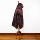 A red and brown patterned poncho drapes over a wooden mannequin in a room with a white wall and wooden floor.