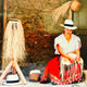 A woman, wearing a white hat, weaves a straw hat. She is seated on a stool, dressed in colorful traditional attire, against an earthy wall backdrop. Nearby, finished hats are displayed.