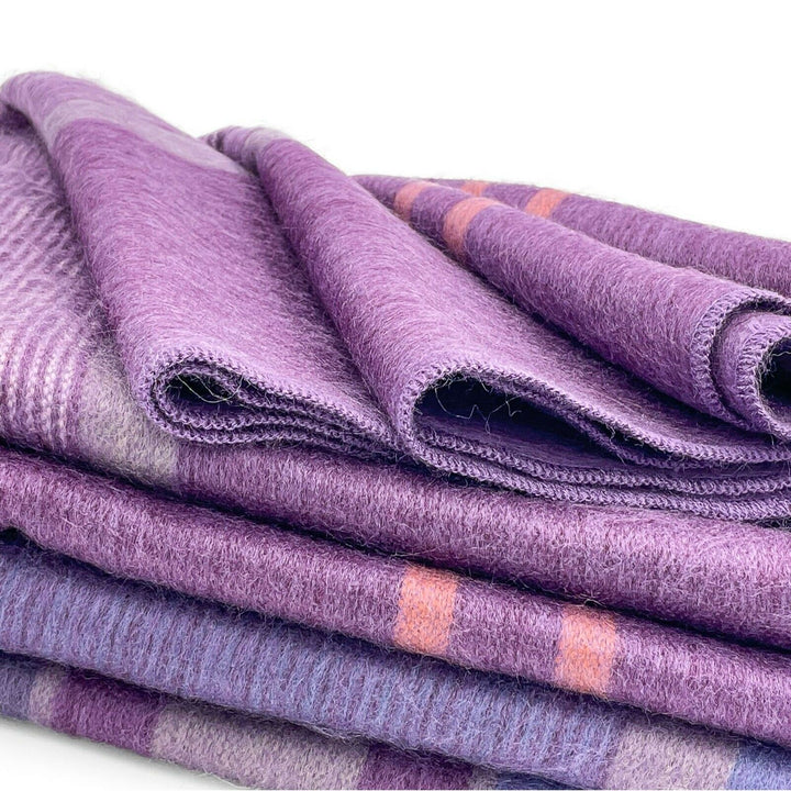 Chuquipata baby alpaca wool blanket – purple woolen blankets are neatly stacked, displaying soft, folded textures with striped and plaid patterns. The blankets are shown in a close-up view, highlighting their fibrous material