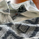 Cuyuja lightweight alpaca wool hooded poncho – a close-up of a woolen poncho with black geometric patterns and fringe details, casually draped on a surface. The background shows a wooden texture