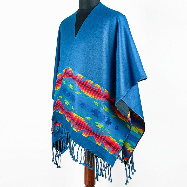 A blue poncho with vibrant geometric patterns and fringe drapes over a mannequin. The design features a colorful, symmetrical band, set against a plain background, suggesting traditional or cultural influences.