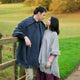 Two people, wrapped in woolen ponchos, stand closely near a wooden fence. They gaze at each other in a grassy field with autumnal trees in the background.