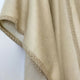 A beige fabric with a textured, embroidered edge drapes softly, creating gentle folds. The backdrop is a plain white wall, highlighting the fabric's smooth texture and intricate border detail.