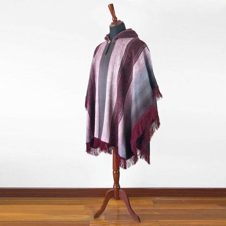 A maroon and gray striped poncho with fringe is displayed on a wooden mannequin stand against a plain white wall and wood floor.