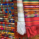 Colorful woven textiles hang, showcasing intricate patterns and fringes. They are stacked vertically in a vibrant display, creating a rich, textured visual in a fabric store setting.