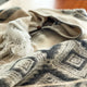 A soft, patterned woolen blanket with geometric designs and fringed edges lies spread out on a wooden surface, suggesting warmth and comfort in a cozy interior setting.