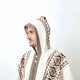 A person, wearing a hooded, cream-colored sweater with geometric brown and black patterns, looks to the side against a plain white background.