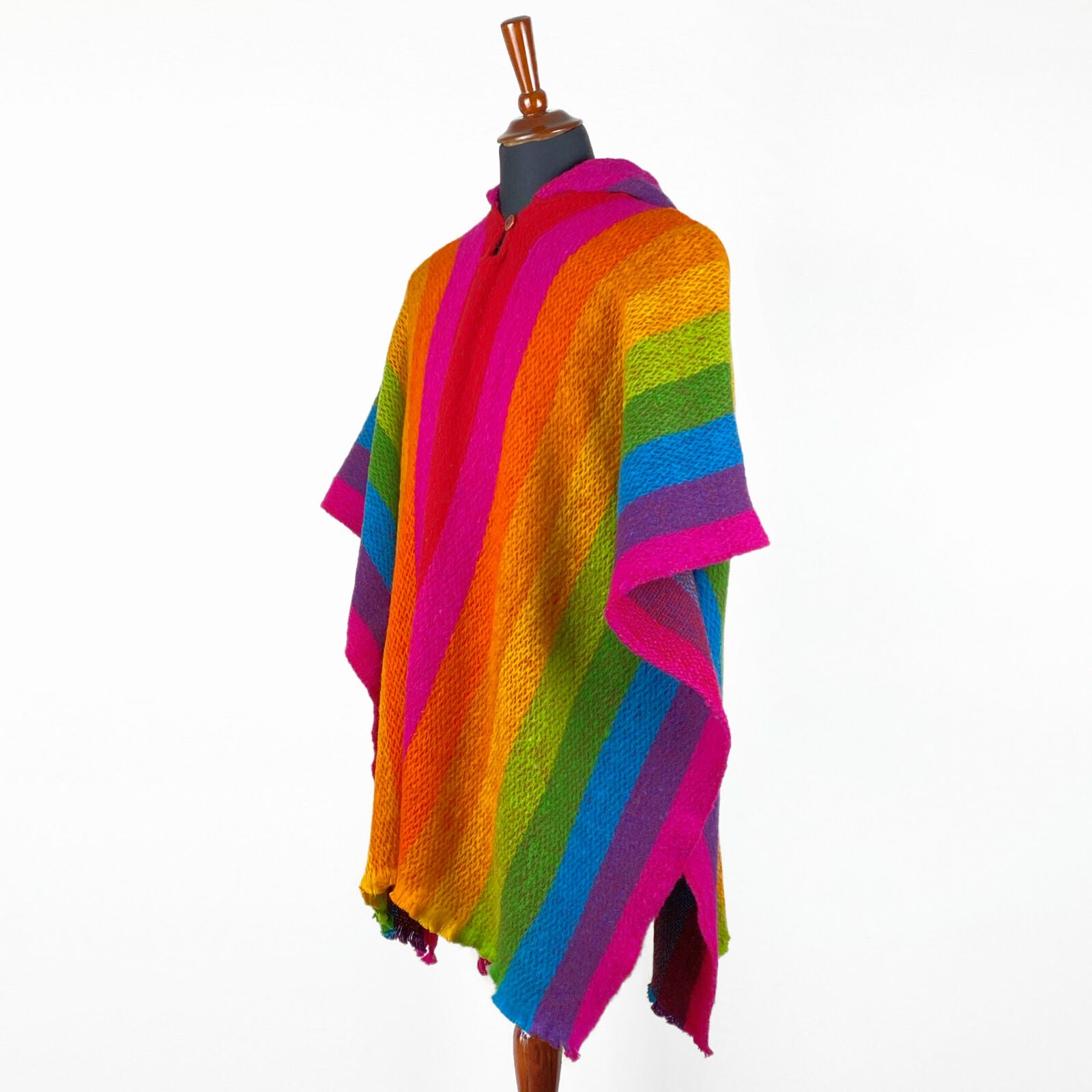 A multicolored poncho with wide vertical stripes in orange, pink, green, blue, and purple hangs on a wooden mannequin against a plain white background.