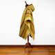 A hooded poncho with intricate geometric patterns and earthy tones is displayed on a wooden stand, positioned within a room with a polished hardwood floor and plain white wall.