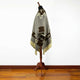 A dark brown and gray poncho with geometric and floral patterns drapes over a wooden mannequin stand on a polished wooden floor against a plain white background.
