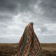 A person draped in a patterned blanket walks across an open, dry landscape under a cloudy sky. The blanket features diamond motifs with orange and brown stripes.