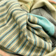 Cojitambo alpaca wool throw blanket – striped fabric with pastel colors of green, blue, yellow, and gray drapes organically, creating folds and waves. The soft texture is highlighted in natural light