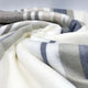 Yacoubina baby alpaca wool blanket – a soft, striped blanket in shades of gray, beige, and white is casually draped and folded, creating a cozy and inviting texture against a plain, light background