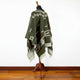 Clint Eastwood alpaca wool poncho – a green poncho with white geometric patterns hangs on a mannequin stand. The scene is set against a plain white wall and wooden floor