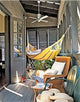 A striped hammock hangs on a cozy porch, surrounded by wicker chairs and a small table with plants, under a ceiling fan. A door opens into the adjacent room.