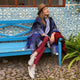 A person sits leisurely on a blue bench, wearing a wide-brimmed hat and colorful clothes, set against a blue patterned wall and cobblestone ground, with greenery nearby.