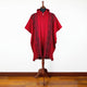 A red poncho with intricate black geometric patterns is displayed on a mannequin. It features a hood and fringe hem, set against a plain white wall on a wooden floor.