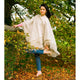 A person wearing a cream poncho and blue jeans balances barefoot on one leg while holding a shoe. They are surrounded by leafy trees and autumn leaves on the ground.